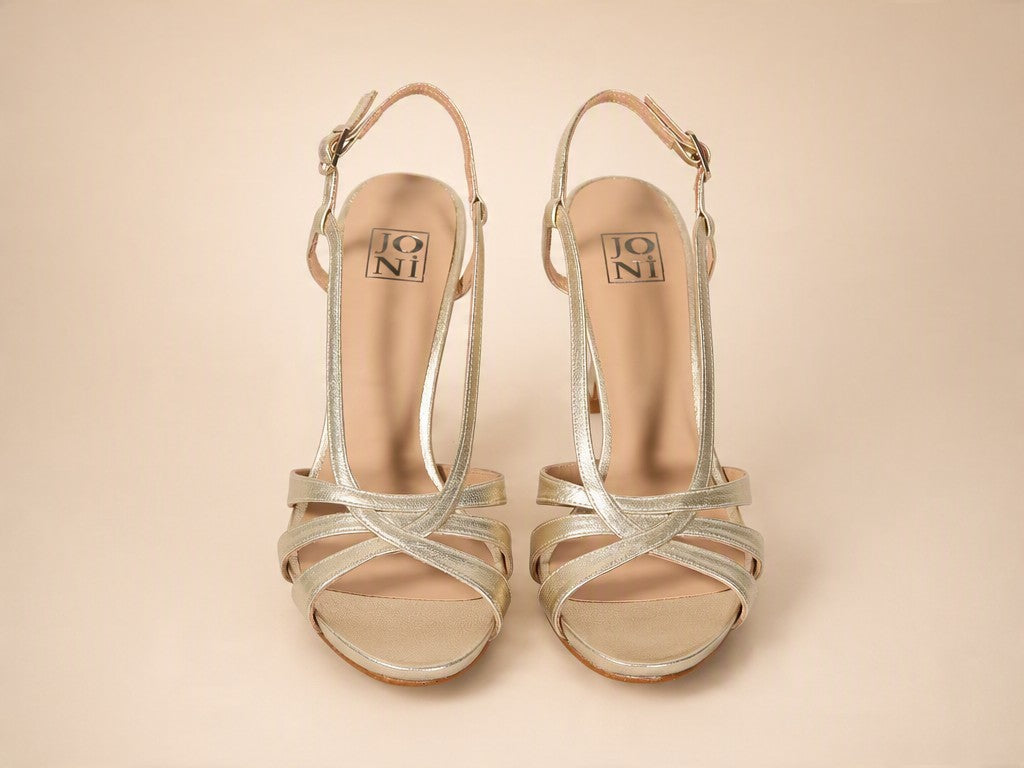 A pair of metallic silver strappy high-heeled sandals with ankle straps, shown from a top-down view on a solid beige background. The brand name 'JONI' is visible on the insole.