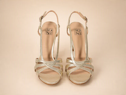 A pair of metallic silver strappy high-heeled sandals with ankle straps, shown from a top-down view on a solid beige background. The brand name 'JONI' is visible on the insole.