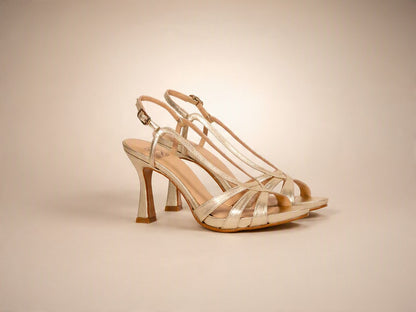 A pair of metallic champagne gold strappy high-heeled sandals with an ankle strap and open toe, shown on a warm beige studio background.